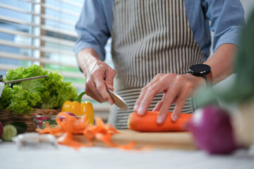 Senior man in apron chopping fresh carrot on board while preparing a healthy salad in kitchen