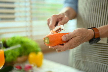 Cropped shot senior man in apron peeling carrot, preparing healthy salad in kitchen