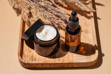 From above open jar of cream with amber glass dropper bottle on a wooden board, pampas grass, beige background