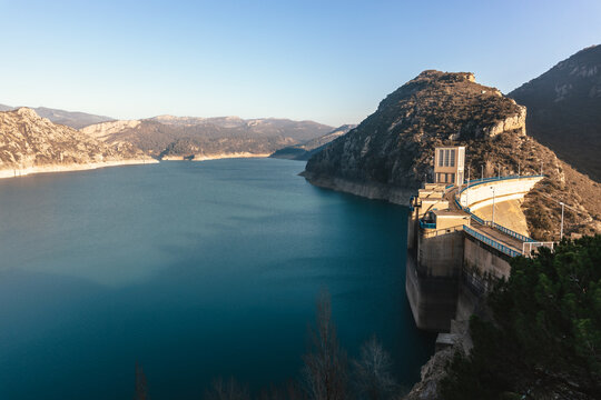 A dam nestled between mountainous terrain with a reservoir displaying low water levels indicative of drought conditions