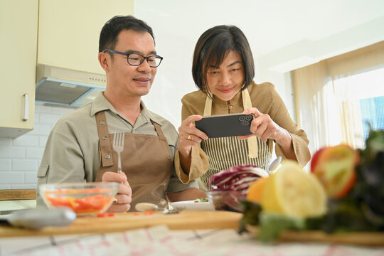 Happy senior woman taking photo of vegan meal with smartphone while cooking with her husband in kitchen.