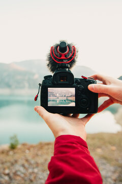 Anonymous hands holding a DSLR camera with a microphone capturing a scenic lake view on the LCD screen