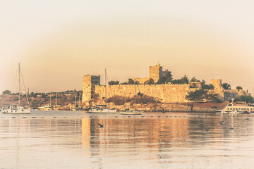 Fototapeta premium Panoramic view to the Bodrum Castle reflecting at waters of harbor at sunrise, flock of birds fluing under calm water. Old postcard stylization. Bodrum (Mugla), Turkey