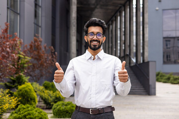 Smiling young man in glasses and white shirt giving thumbs up outside modern office building,...