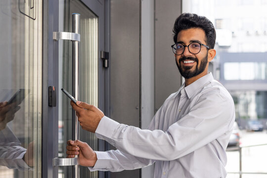 Confident young professional opening office door with smartphone, showcasing modern access technology and a welcoming smile.