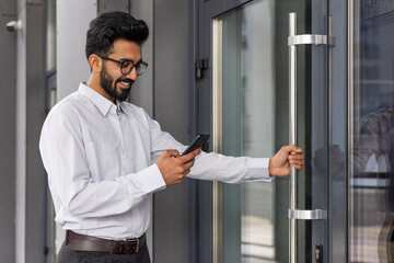 Smiling businessman in glasses using smartphone by office door. Concept of tech-savvy professional, modern communication.