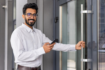 Naklejka na ściany i meble Confident businessman with glasses smiling while holding a smartphone and opening a glass door, depicting connectivity and success.