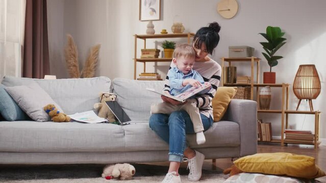 Mother spending time together with little son. Mother reading book to child. Mother reading tales to toddler. Woman enjoying being mother. Little boy attentively looking at pictures in book.