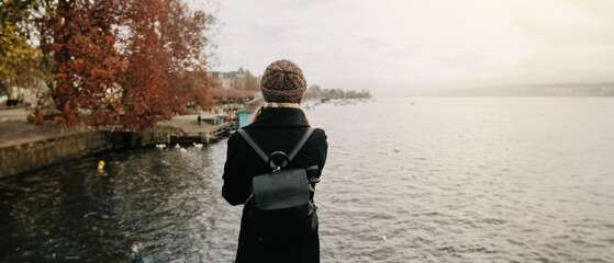 Young beautiful pretty blonde tourist girl in warm hat and coat with backpack walking at cold autumn in Europe city enjoying her travel in Zurich Switzerland