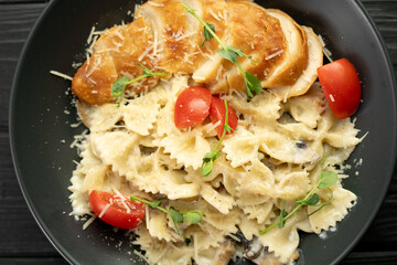 Tasty appetizing classic italian farfalle pasta with chicken breast, cheese parmesan on black plate on dark wooden table. View from above.