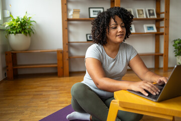 Young woman using laptop while sitting on yoga mat at home - obrazy, fototapety, plakaty