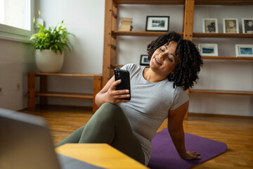 Happy woman using smartphone while sitting on yoga mat at home