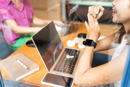 Cropped unrecognizable smiling woman wearing a smartwatch engaging in a conversation with a colleague at a cafe while working on her laptop remote work in Chiang Mai, Thailand