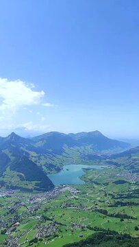 Vertical video panning over a valley with a city, a lake and mountains. Vertical video pan on top of a mountain overlooking a valley with a lake, a city and mountains in the background. Sunny day