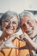 Close up portrait happy sincere middle aged elderly retired family couple making heart gesture with fingers, showing love or demonstrating sincere feelings together indoors, looking at camera..