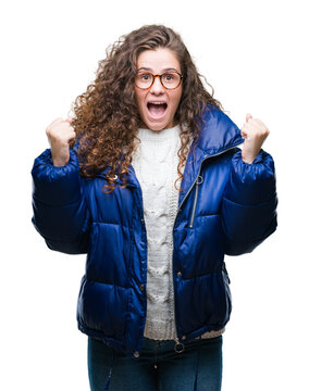 Beautiful young brunette curly hair girl wearing winter coat, glasses and sweater over isolated background celebrating surprised and amazed for success with arms raised and open eyes. Winner concept.