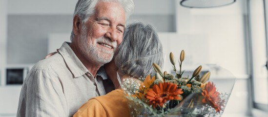 Old man giving flowers at his wife sitting on the sofa at home for the San Valentines’ day....