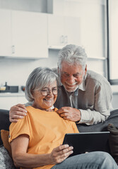 Happy older couple using computer tablet together at home, excited mature man and woman looking at mobile device screen, shopping or chatting online, sitting on cozy couch in living room. © Daniel