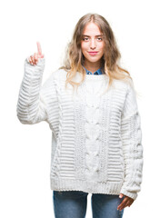 Beautiful young blonde woman wearing winter sweater over isolated background showing and pointing up with finger number one while smiling confident and happy.