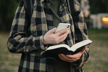A man with a smartphone and a Bible in his hands outside, christian online technology concept, online live church, Holy Bible book and online study. - obrazy, fototapety, plakaty