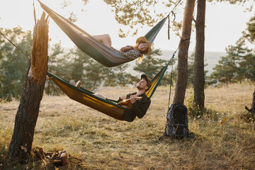 A guy and a girl are relaxing in hammocks in a pine forest at sunset. A couple in love rest in hammocks after a hike in the mountains, a guy plays the ukulele. - obrazy, fototapety, plakaty