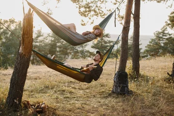 Foto op Plexiglas A guy and a girl are relaxing in hammocks in a pine forest at sunset. A couple in love rest in hammocks after a hike in the mountains, a guy plays the ukulele. © dsheremeta
