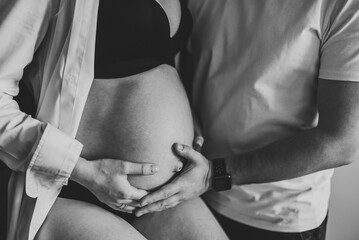 Loving couple. Black and white photo pregnant woman, loving handsome man hugging tummy at home. Waiting baby. Nine months closeup. Husband holding hands on belly of pregnant wife. Parenthood concept.