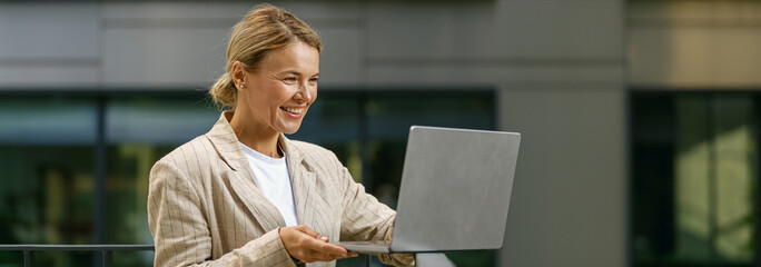 Stylish smiling woman entrepreneur working on laptop outside on modern building background