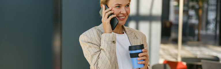 Smiling business woman holding coffee and talking phone standing on modern office background