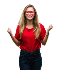 Young beautiful blonde woman wearing glasses over isolated background celebrating surprised and amazed for success with arms raised and open eyes. Winner concept.