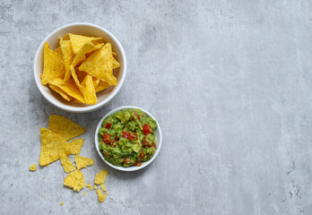 Top view of tortilla chips in a white bowl and avocado guacamole on grey table 