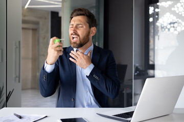A young male businessman suffers from a severe sore throat, sits at a desk in the office and snorts a medicine aerosol into his mouth - obrazy, fototapety, plakaty