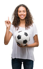 Young hispanic woman holding soccer football ball surprised with an idea or question pointing finger with happy face, number one