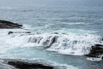 waves crashing against dark rocky shores, creating white foam, under a cloudy sky, showcasing nature s powerful yet serene beauty