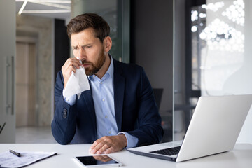 Naklejka premium A sick young man businessman sits in the office at a desk with a laptop and documents, wipes his nose with a napkin, suffers from a cold and a virus