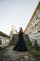 Woman in black long dress walk on stairs near ancient palace at sunset. Model girl goes in street. Luxury bride near old Pidhirtsi Castle, Lviv, Ukraine. Stylish female outdoors. Back view.