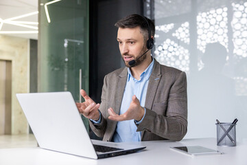 Focused businessman with headset engaged in online communication at modern office, gesturing at laptop screen