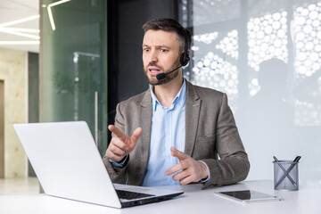 Professional man in headset speaking gesturing at laptop, office setting, customer service, online helpdesk, remote communication concept.