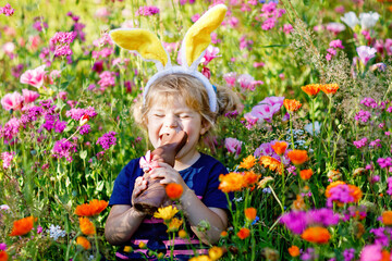 Portriat of adorable, charming toddler girl with Easter bunny ears eating chocolate bunny figure in...