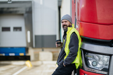 Truck driver leaning against red truck and drinking coffee, waiting for warehouse workers.