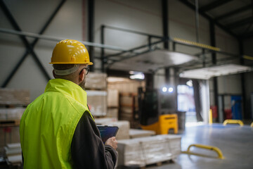 Warehouse receiver standing by delivered cargo, holding tablet, looking at cargo details, checking...