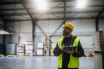 Warehouse receiver standing by delivered cargo, holding tablet, looking at cargo details, checking...