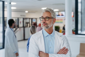 Portrait of confident mature doctor standing in Hospital corridor. Handsome doctor with gray hair wearing white coat, stethoscope around neck standing in modern private clinic, looking at camera. - obrazy, fototapety, plakaty