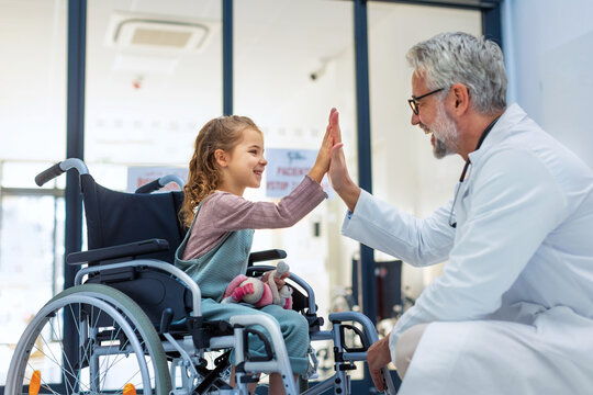 Friendly pediatrician giving high five to little patient in wheelchair. Cute preschool girl in wheelchair greeting doctor in hospital.