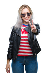 Young blonde woman wearing fashion jacket and sunglasses over isolated background smiling with happy face winking at the camera doing victory sign. Number two.