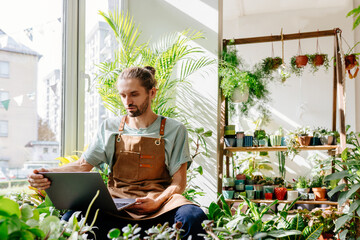 Small business, home decor concept. Handsome caucasian man owner of small business flower shop using technology to streamline daily flower shop related tasks uses computer comfortably on windowsill. - obrazy, fototapety, plakaty