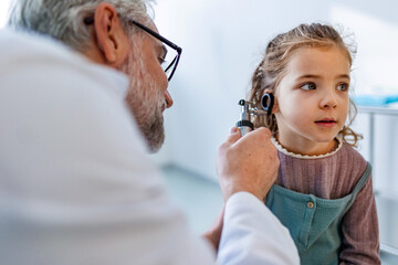 Doctor examining little girl's ear using otoscope, looking for infection. Friendly relationship between the doctor and the child patient. - obrazy, fototapety, plakaty
