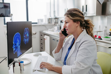 Female doctor working on computer in doctor's office, looking at MRI scan, phone calling test...