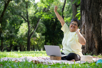 Happy Asian senior woman using laptop for working online outside office. Elderly retirement using laptop for learning new skill and freelance working in park, senior lifestyle with technology