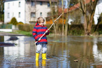 Little kid boy wearing yellow rain boots and walking and jumping into puddle on warm sunny spring day.